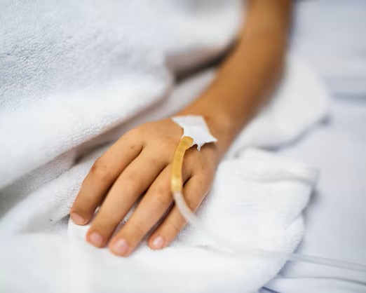 Close-up of a child or young person's hand resting on a white towel or blanket in a hospital bed, with an intravenous (IV) catheter inserted and secured with tape.