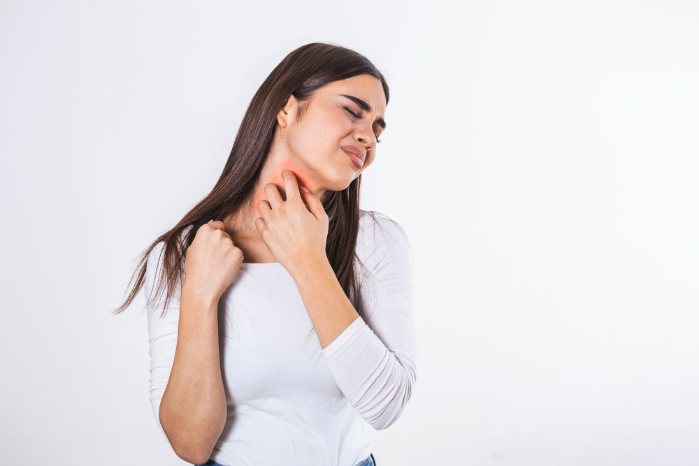 A woman in a white shirt scratches the side of her neck where a prominent red rash or irritated patch of skin is visible, indicating severe itching or an allergic reaction.