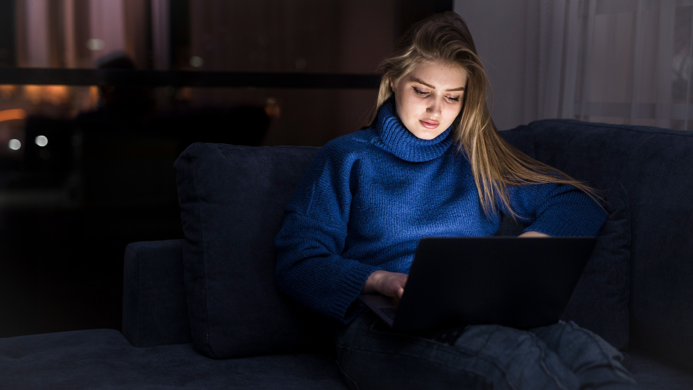 A young woman wearing a blue turtleneck sweater sits on a couch in a dark room at night, illuminated only by the screen of the laptop she is working on.
